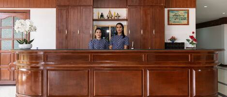 A reception desk with two individuals standing behind it, wooden paneling, a chandelier, and a painting on the wall.
