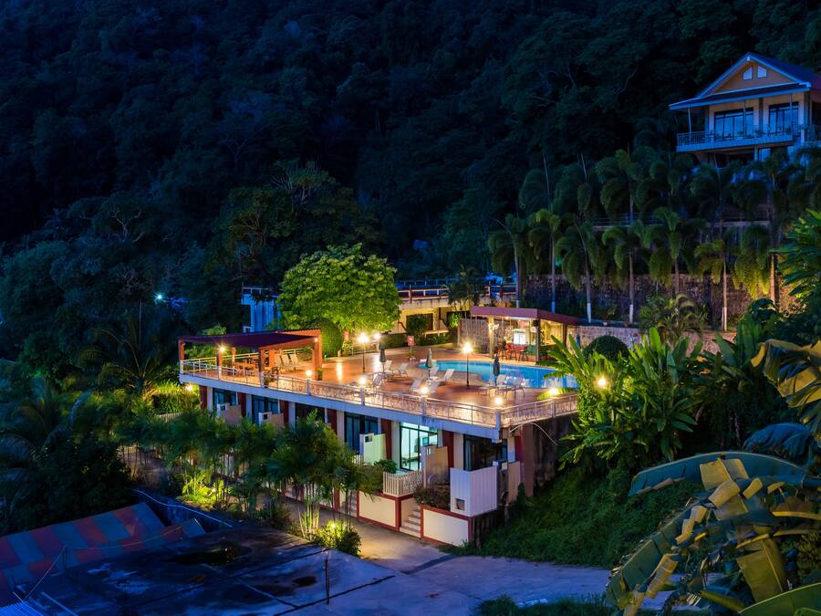 Un hotel de varios pisos con piscina, rodeado de abundante vegetación y palmeras.