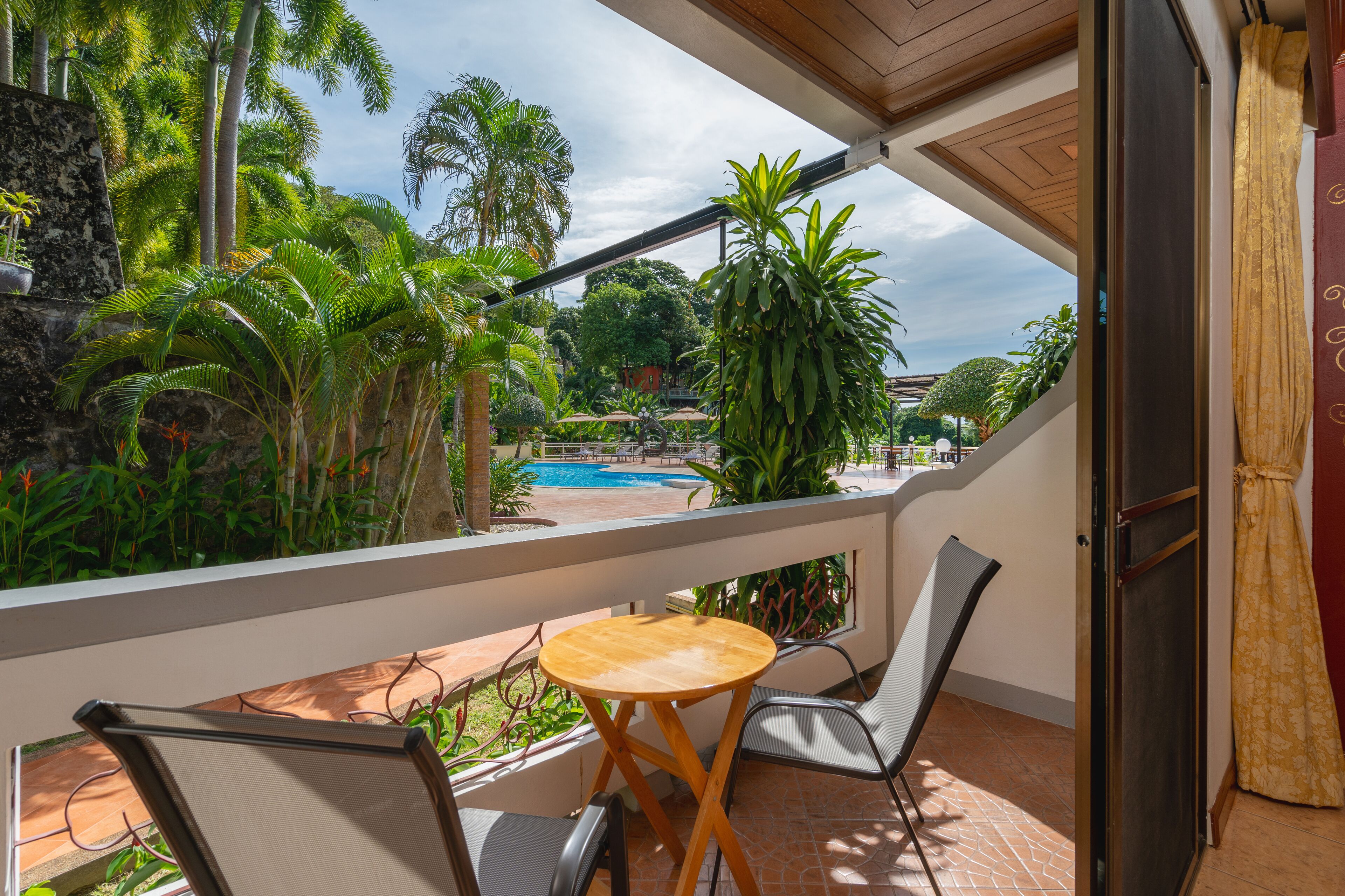 Ein Balkon mit Blick auf ein Schwimmbecken, umgeben von üppiger Bepflanzung und einem roten Gebäude im Hintergrund.