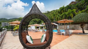 A wicker swing chair with brown cushions and a decorative top, a pool area with lounge chairs and umbrellas, and a mountainous backdrop. - Memory Karon Resort (Karon)