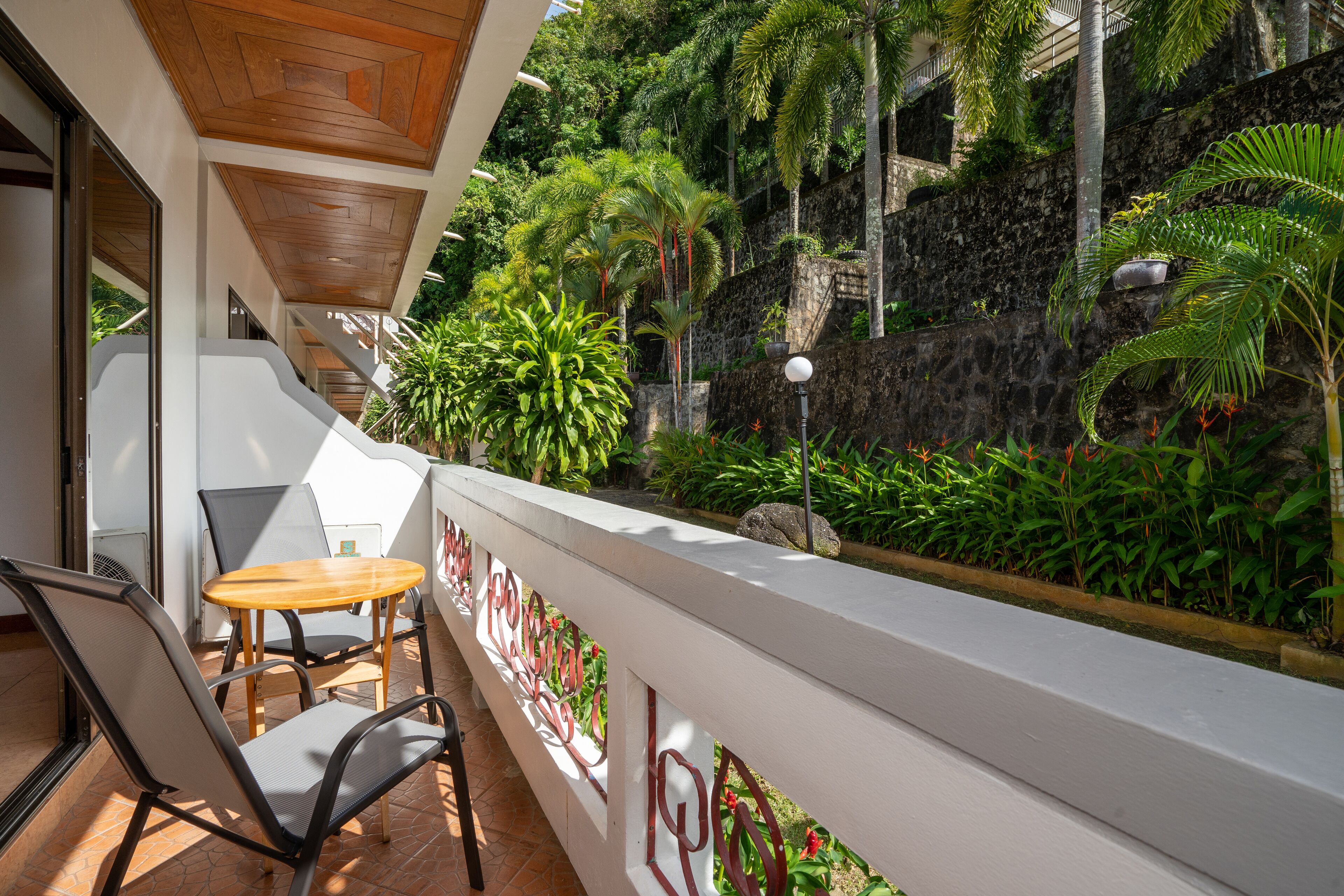 A balcony with a table and chairs, surrounded by lush greenery and a stone wall.