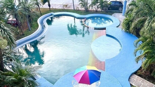 Piscina com design ondulado, cercada por palmeiras e área de deck azul com mesa e guarda-sol.