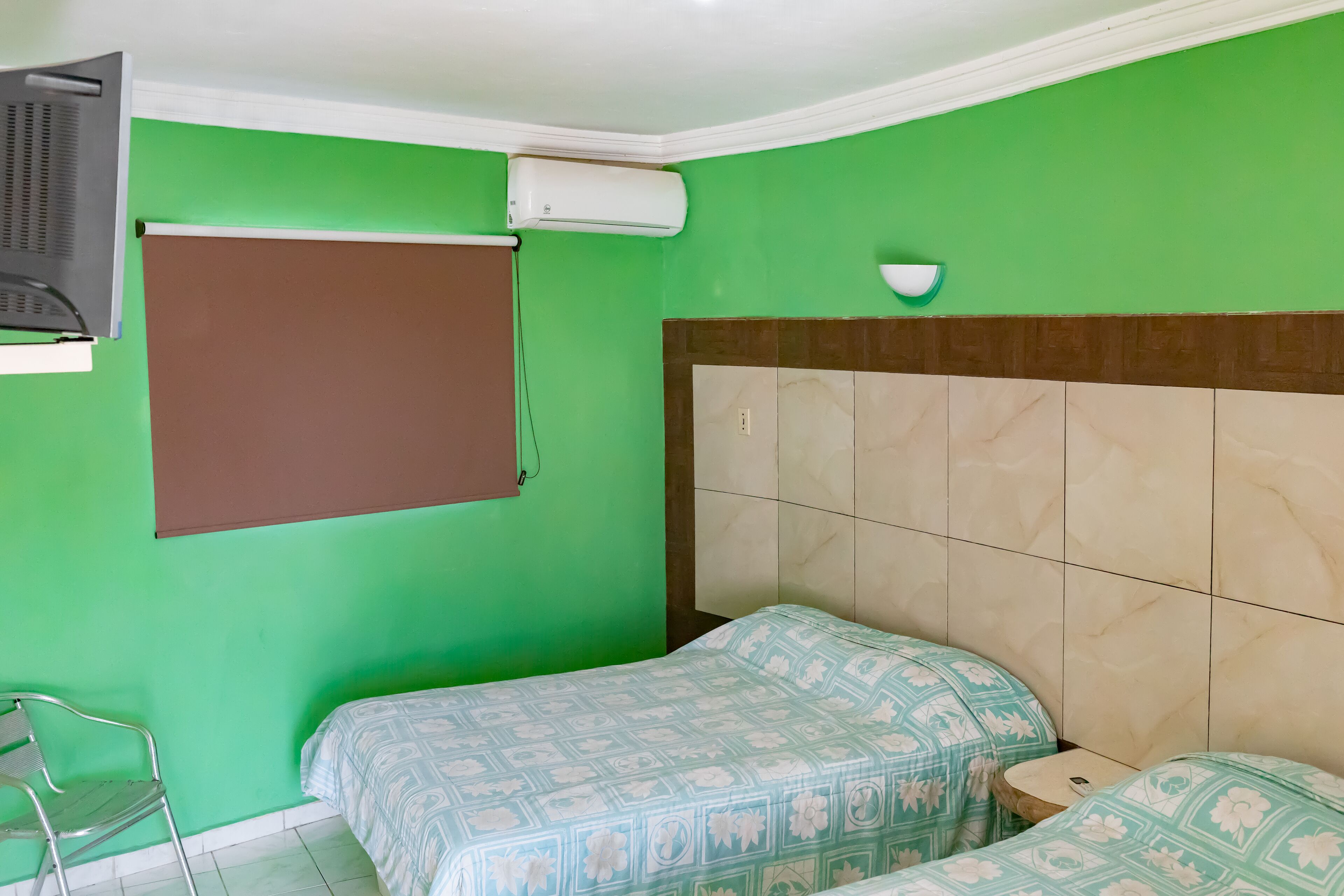 A room with green walls, a brown window blind, a white air conditioner, a small bedside table, and a bed with floral-patterned bedding.