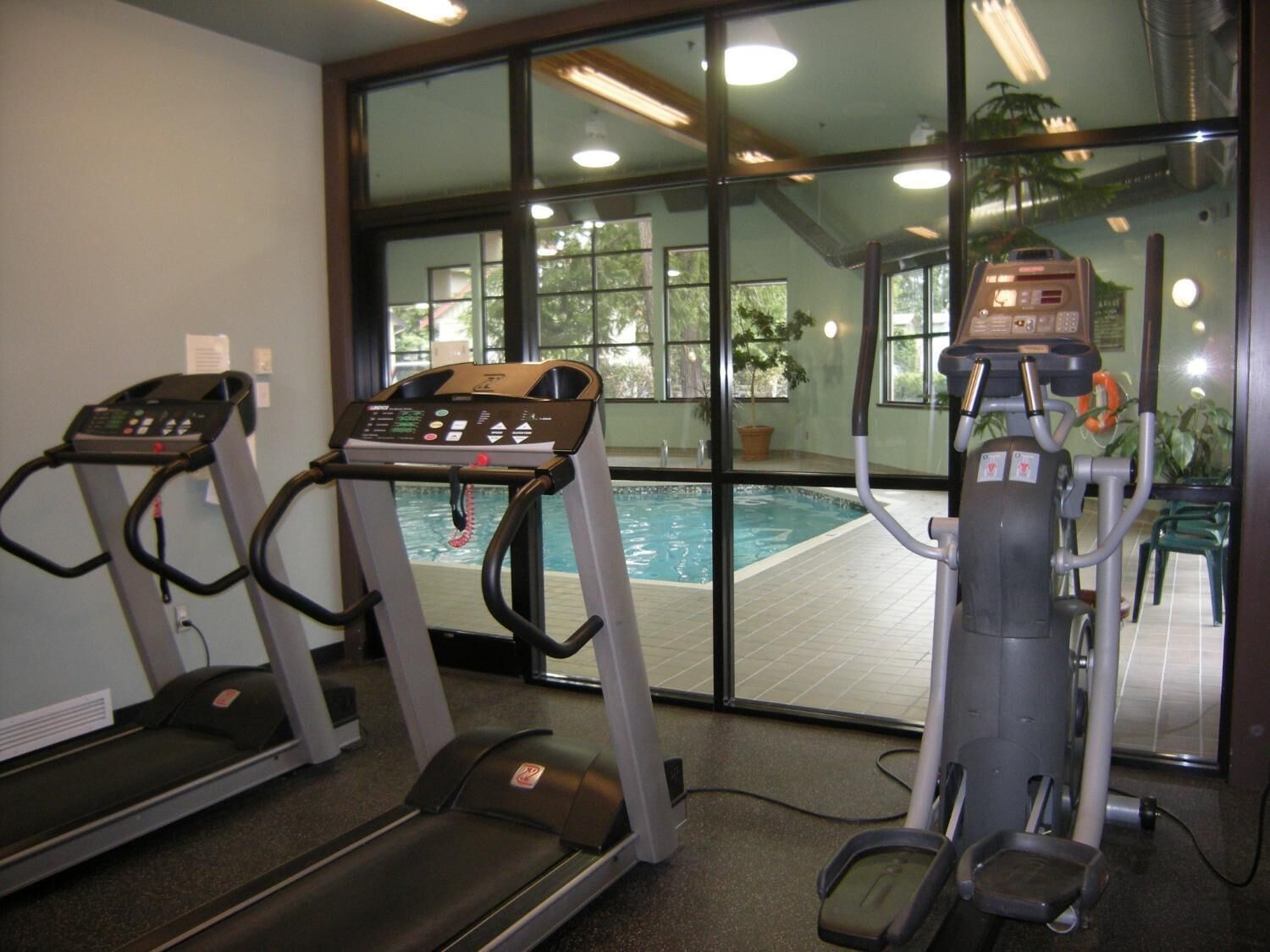 Et fitnessrum med løbebånd, crosstrainer og et poolområde, alt sammen synligt gennem store vinduer.