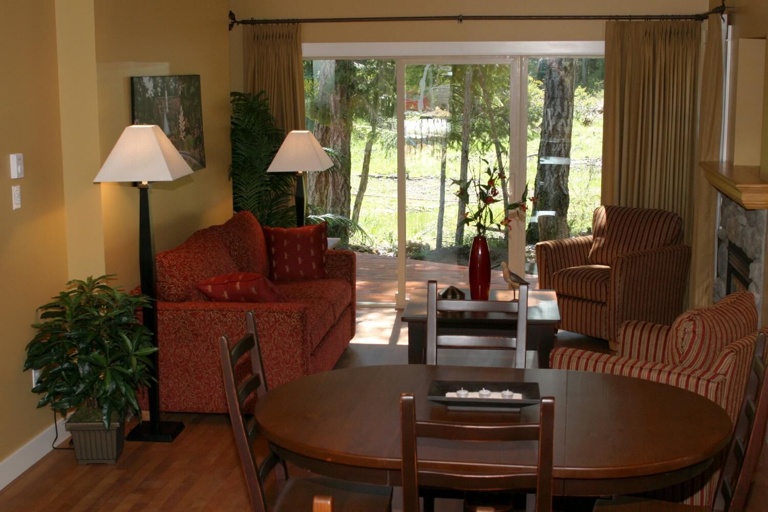A cozy living room with a red sofa, striped armchairs, a dining table with chairs, two floor lamps, and a large window with curtains.