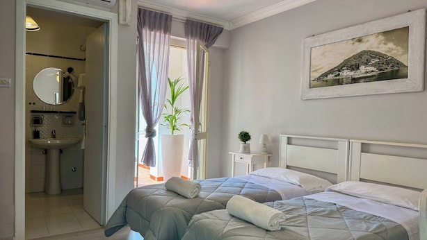Une chambre avec deux lits, une table de chevet, une commode, une grande fenêtre avec des rideaux, une salle de bain avec un lavabo et un miroir, et un tableau encadré au mur.