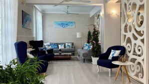 Un salon moderne avec un canapé gris, des fauteuils bleus, une table basse et des coussins décoratifs. On y trouve également un ventilateur de plafond blanc, un grand tableau abstrait et un élément de séparation décoratif blanc orné de motifs complexes.