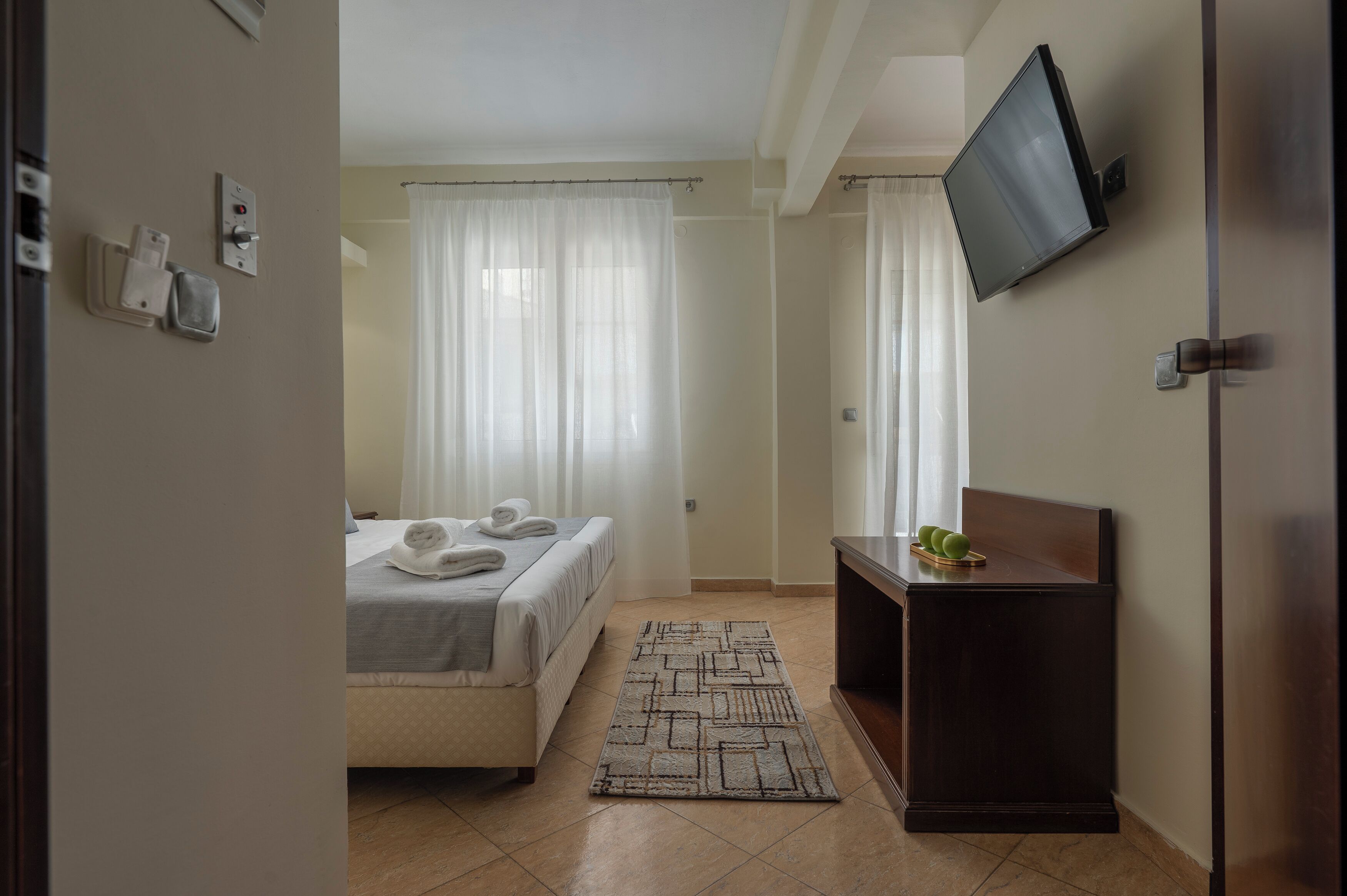 Une chambre d’hôtel avec un lit, un téléviseur fixé au mur, une table de chevet avec un panier de fruits et un tapis à motifs.