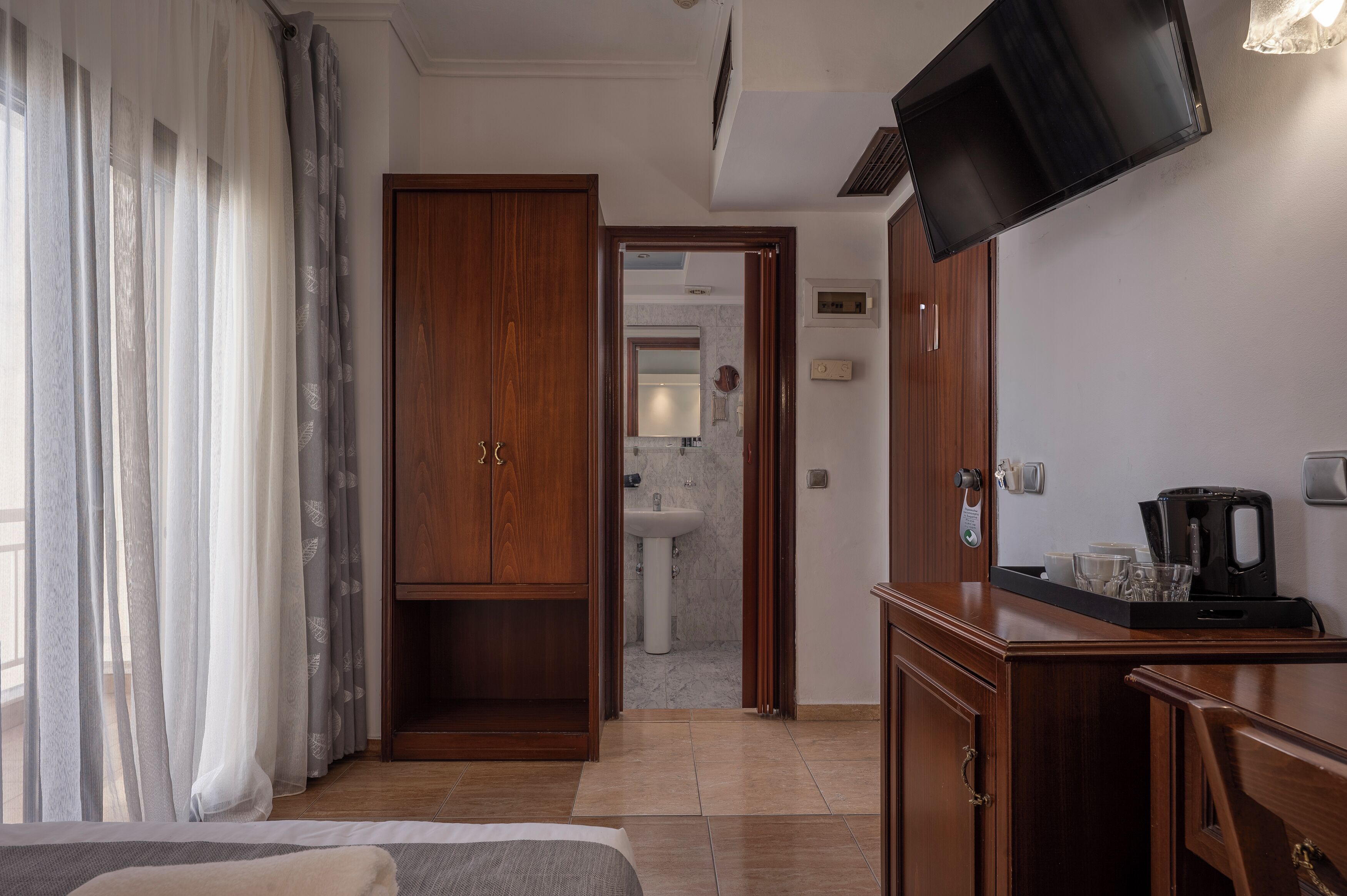 Une chambre d’hôtel avec un lit, une armoire en bois, un téléviseur fixé au mur, une machine à café et une salle de bain visible à travers une porte ouverte.