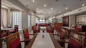 A spacious dining area with multiple tables and chairs, a kitchen area, and large windows allowing natural light.