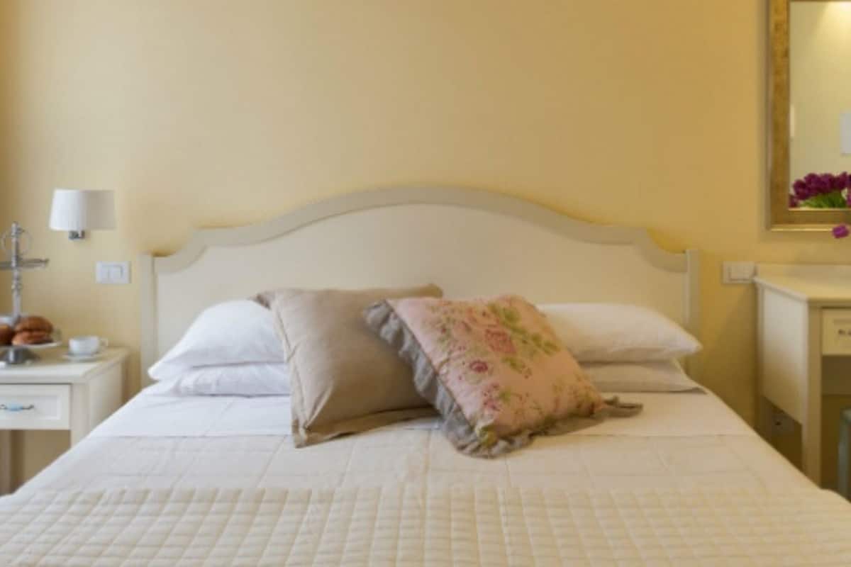 A neatly made bed with a quilted cover, a bedside table with a lamp, a chair, and a desk with a lamp and flowers.