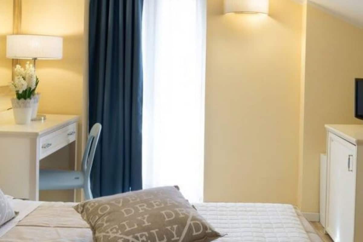 A hotel room with a bed, a desk with a lamp, a television, and a window with curtains.