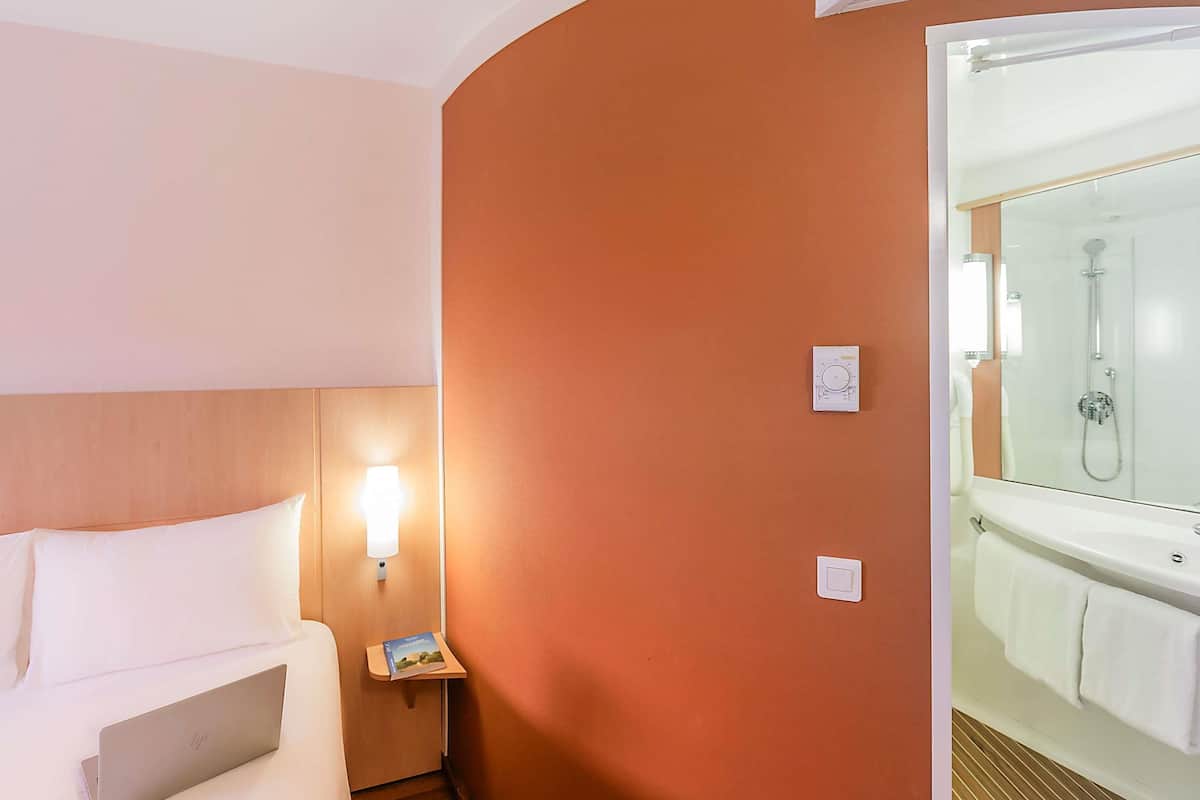 A hotel room with a bed, a nightstand, a reading light, a wall-mounted thermostat, and a glass-enclosed shower area.
