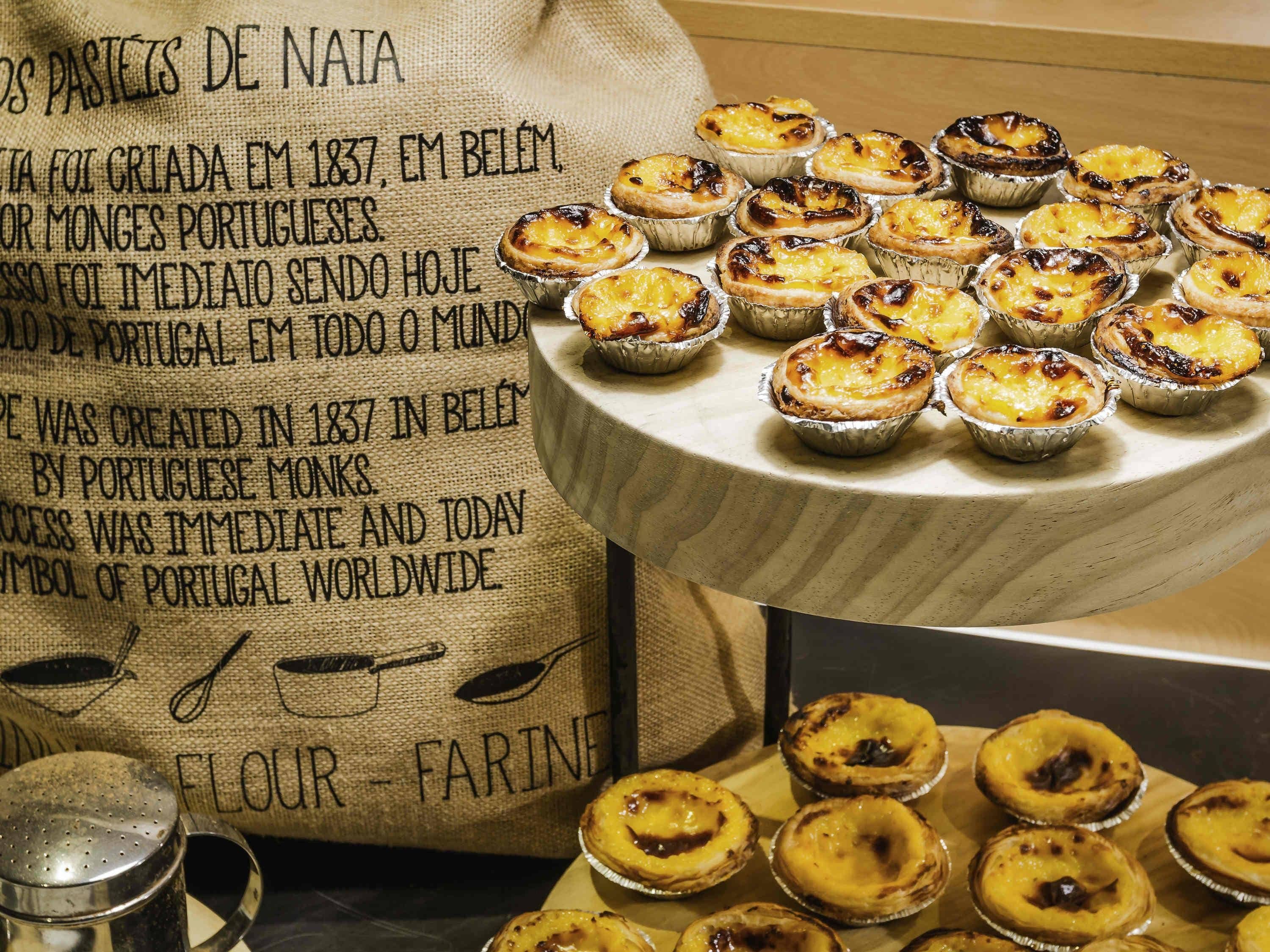 Una mesa con varios pasteles portugueses de crema y un saco de arpillera con texto.