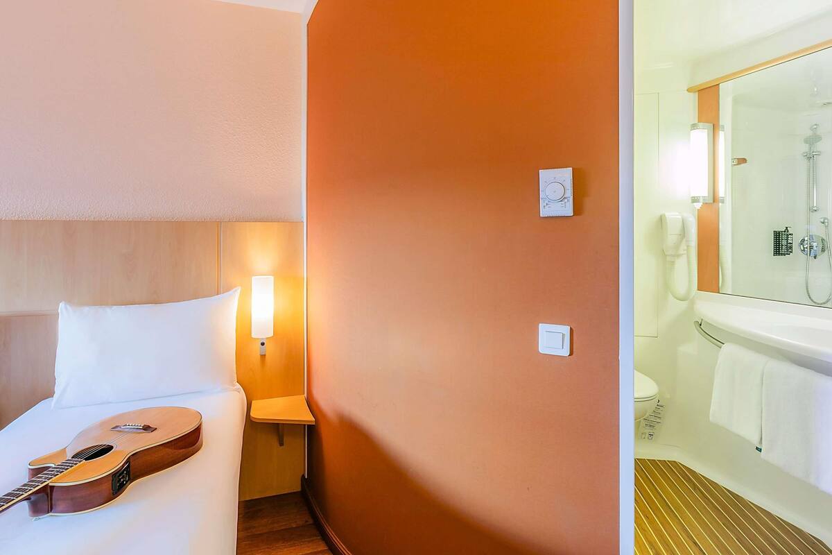 A hotel room with a bed, a guitar, a nightstand, a wall-mounted lamp, a thermostat, and a bathroom with a bathtub and a mirror.