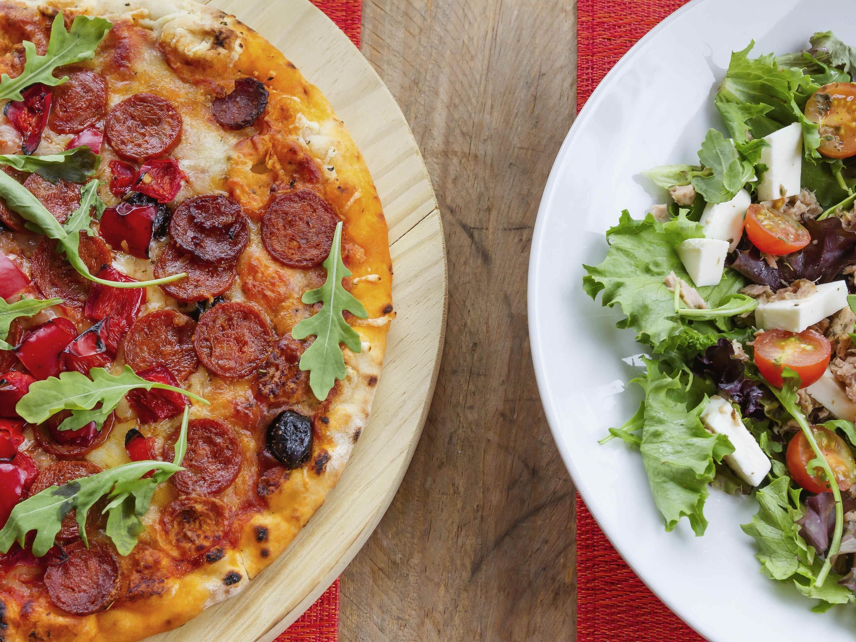 Pizza con pepperoni y rúcula sobre una tabla de madera, junto a un plato con ensalada.