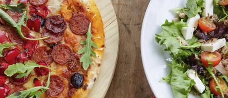 Pizza con pepperoni y rúcula sobre una tabla de madera, junto a un plato con ensalada.