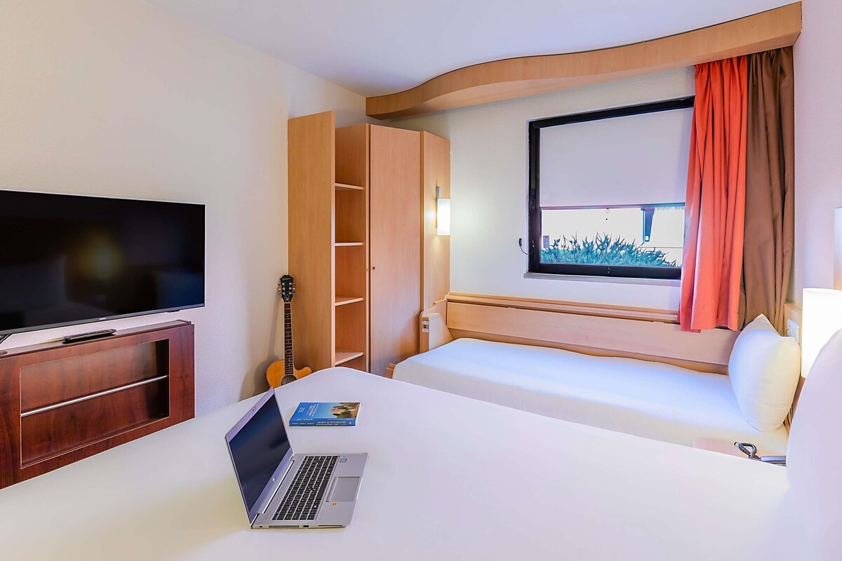 A hotel room with a bed, a TV, a laptop, a guitar, and a window with curtains.