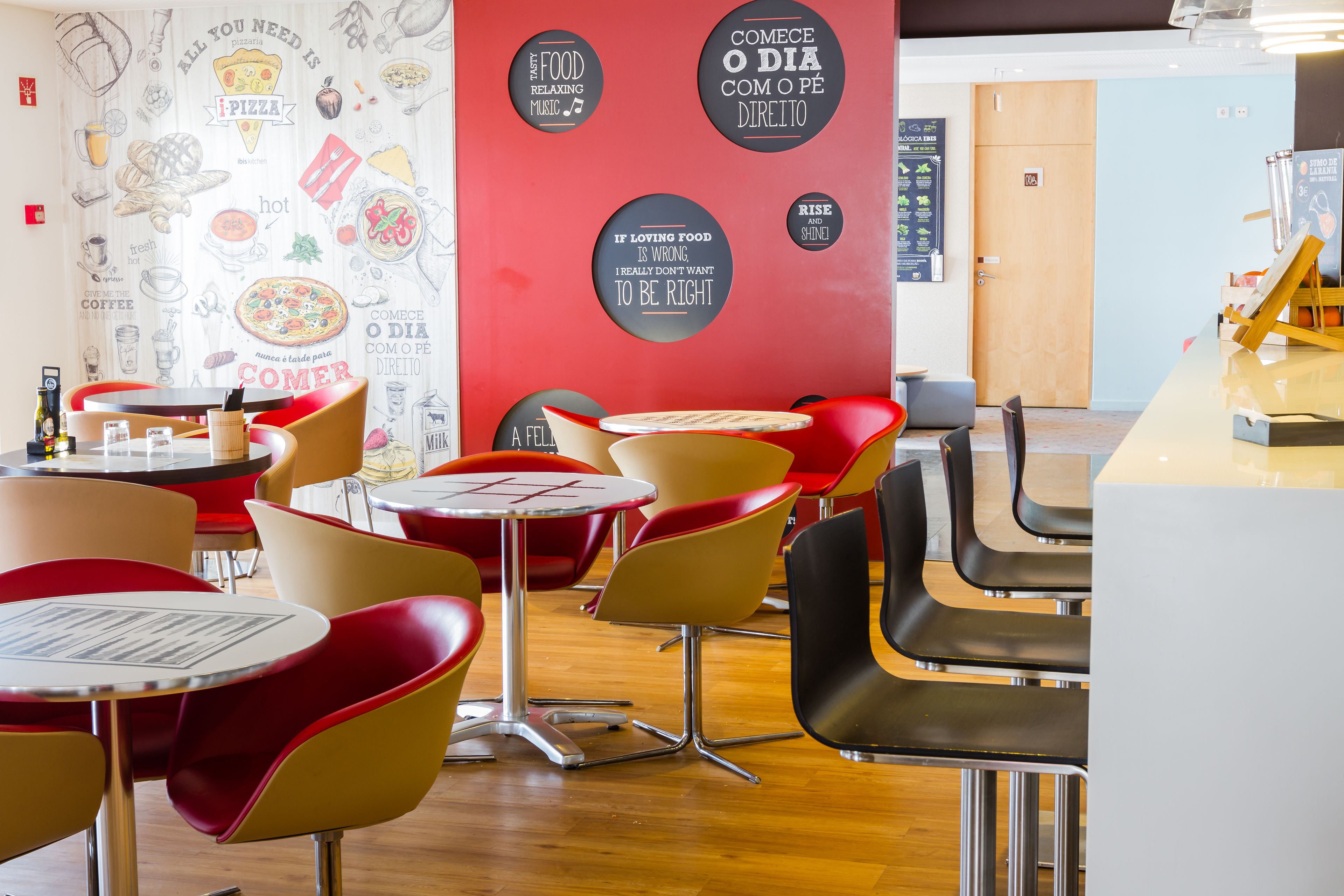 A modern cafe interior with red and yellow chairs, a wooden floor, and a wall decorated with food-related illustrations and text.