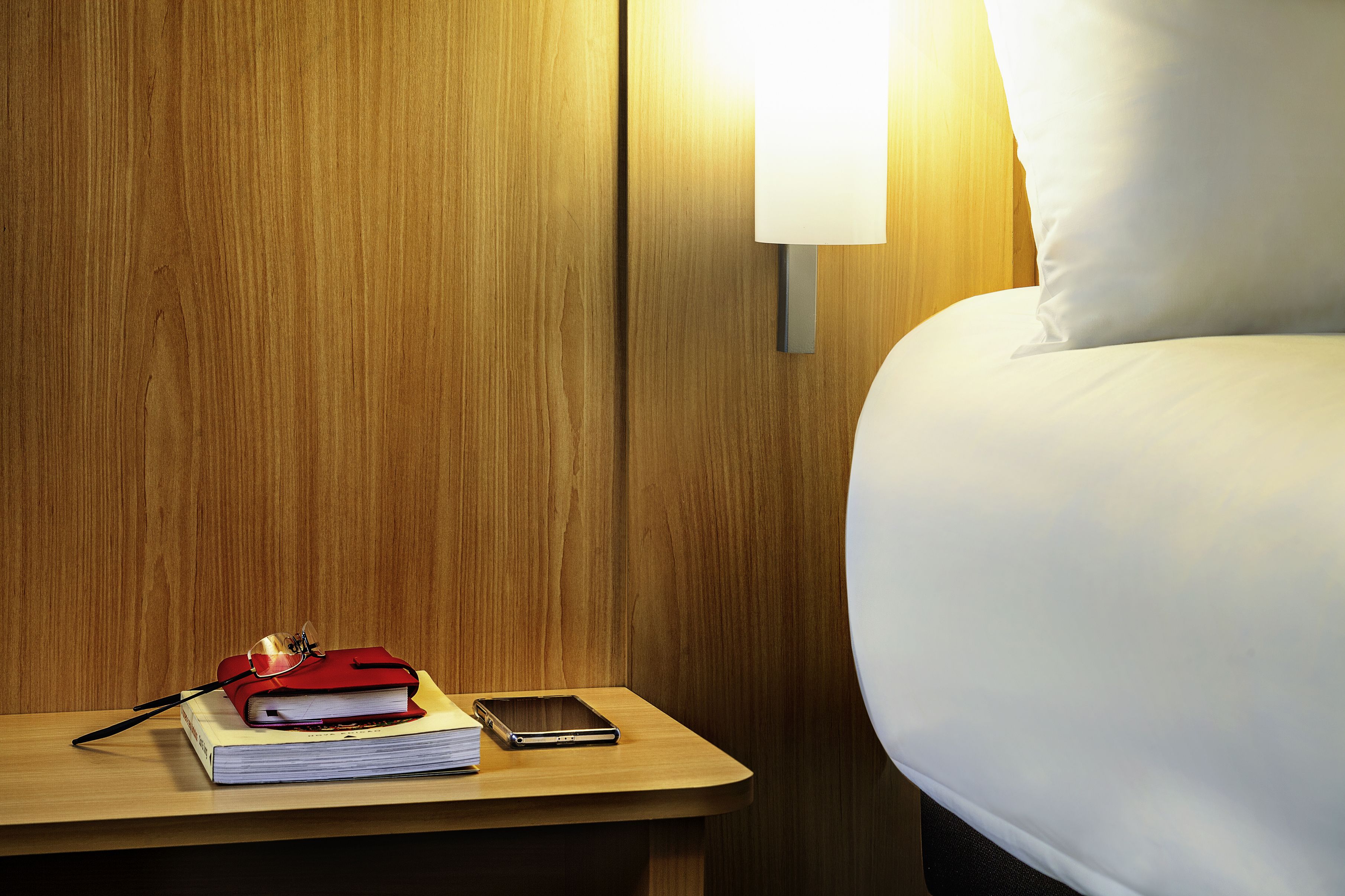 Una cama bien hecha con un almohadón blanco y una mesita de noche de madera. Sobre la mesita, un libro rojo, un teléfono inteligente y un par de gafas.