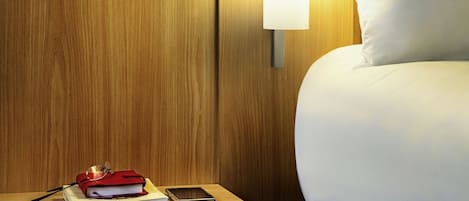 A neatly made bed with a white pillow and a wooden nightstand beside it. On the nightstand, there is a red book, a smartphone, and a pair of glasses.