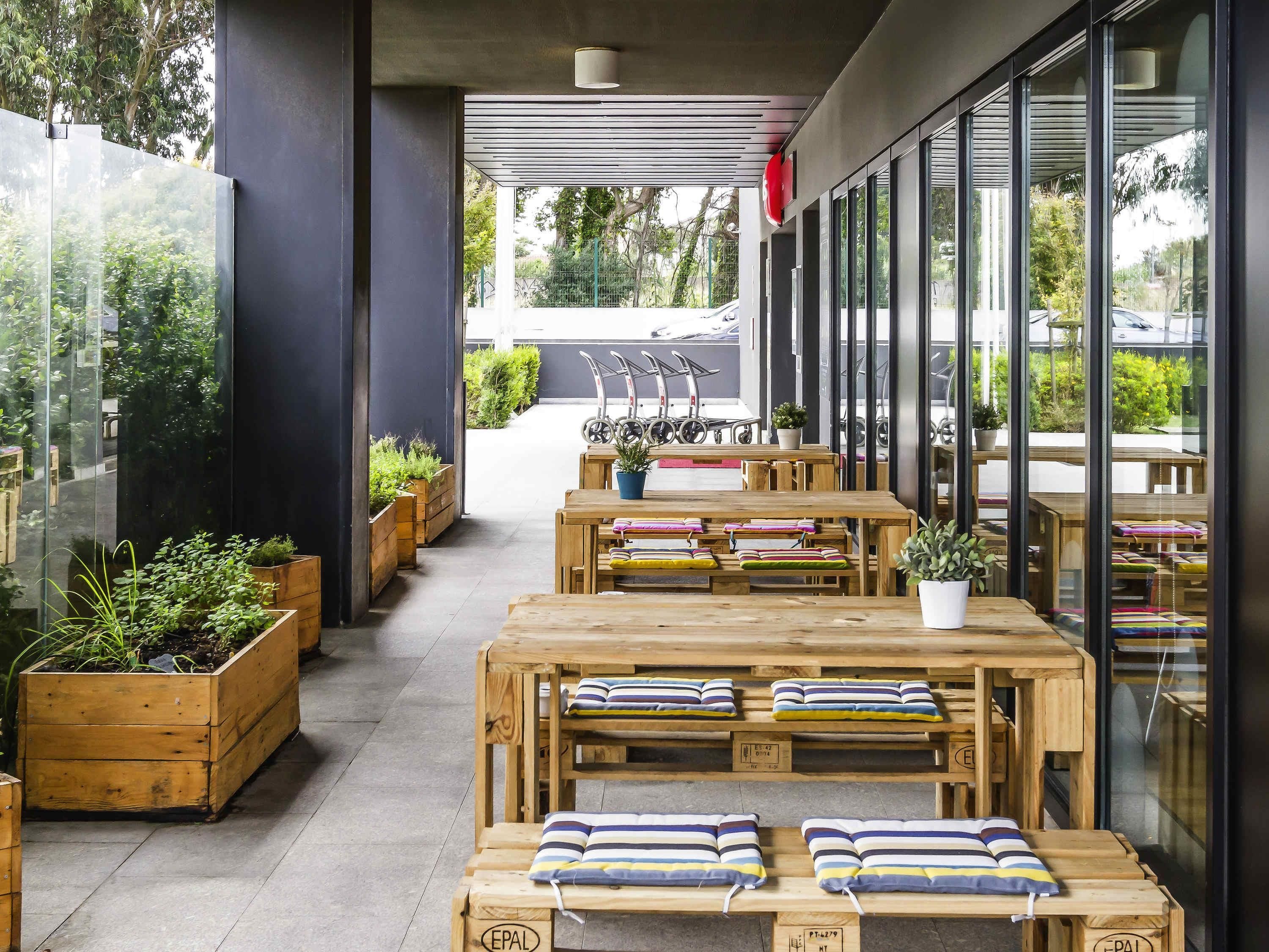 Un área de estar al aire libre con mesas y bancos de madera, plantas en macetas y vistas a la calle.
