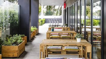Un área de estar al aire libre con mesas y bancos de madera, plantas en macetas y vistas a la calle.