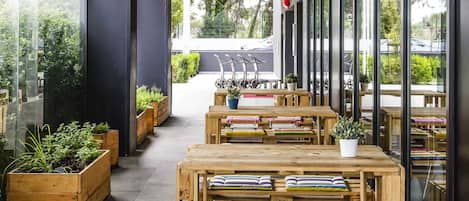 Un área de estar al aire libre con mesas y bancos de madera, plantas en macetas y vistas a la calle.