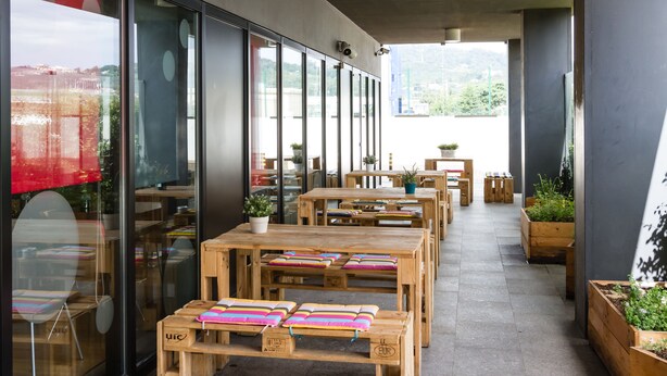 Een terras met houten tafels en banken, plantenbakken en uitzicht op de stad door grote ramen.