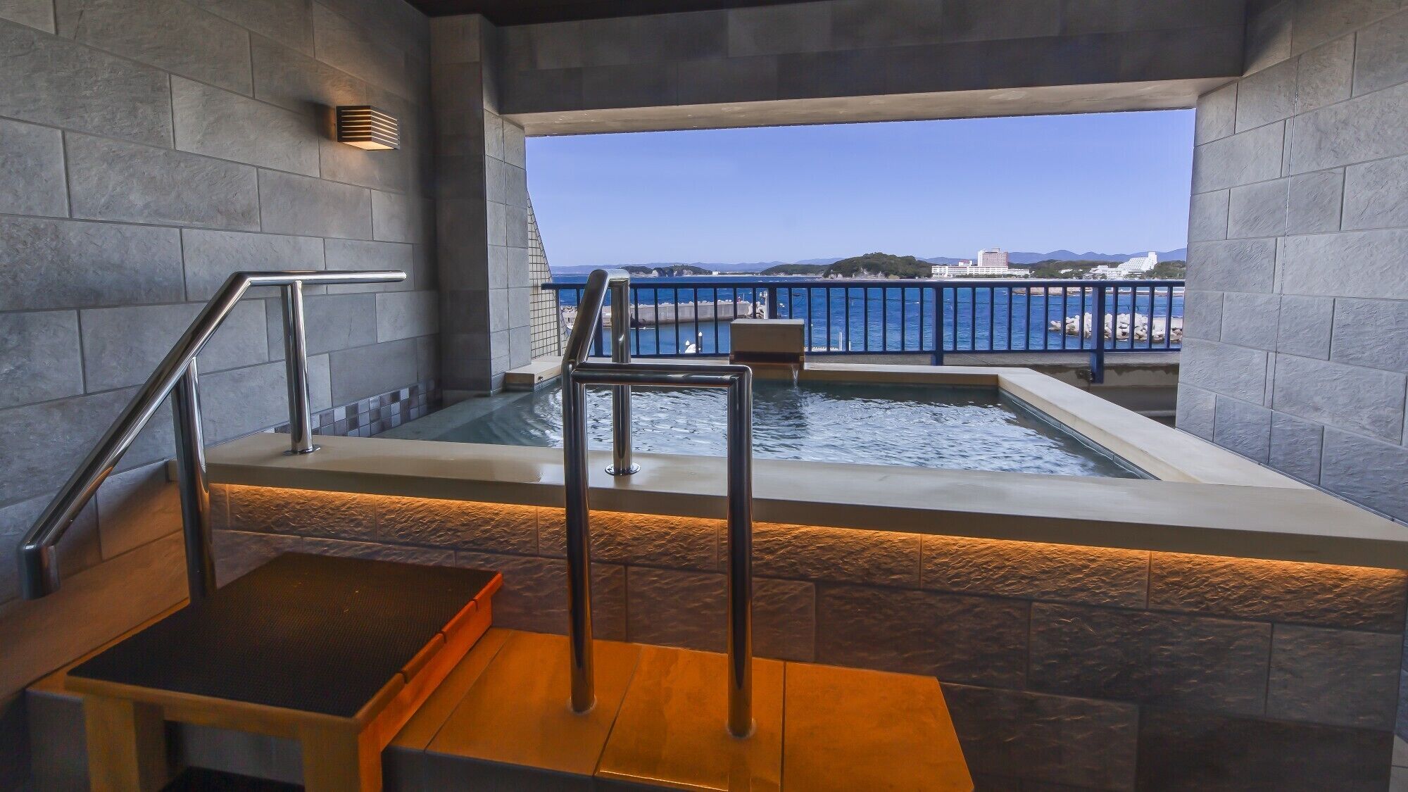 A modern indoor pool with a view of the sea, a metal staircase, and a stone wall.