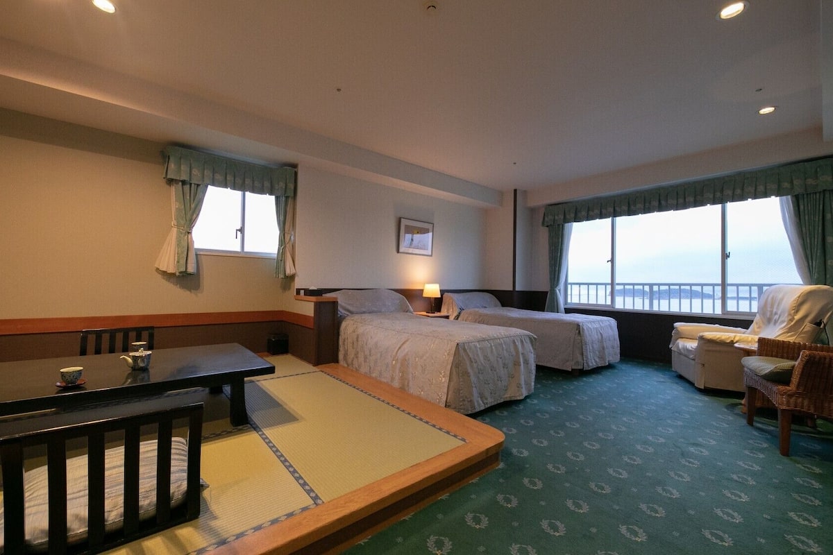 A hotel room with two beds, a dining table, a chair, and a view of the sea.