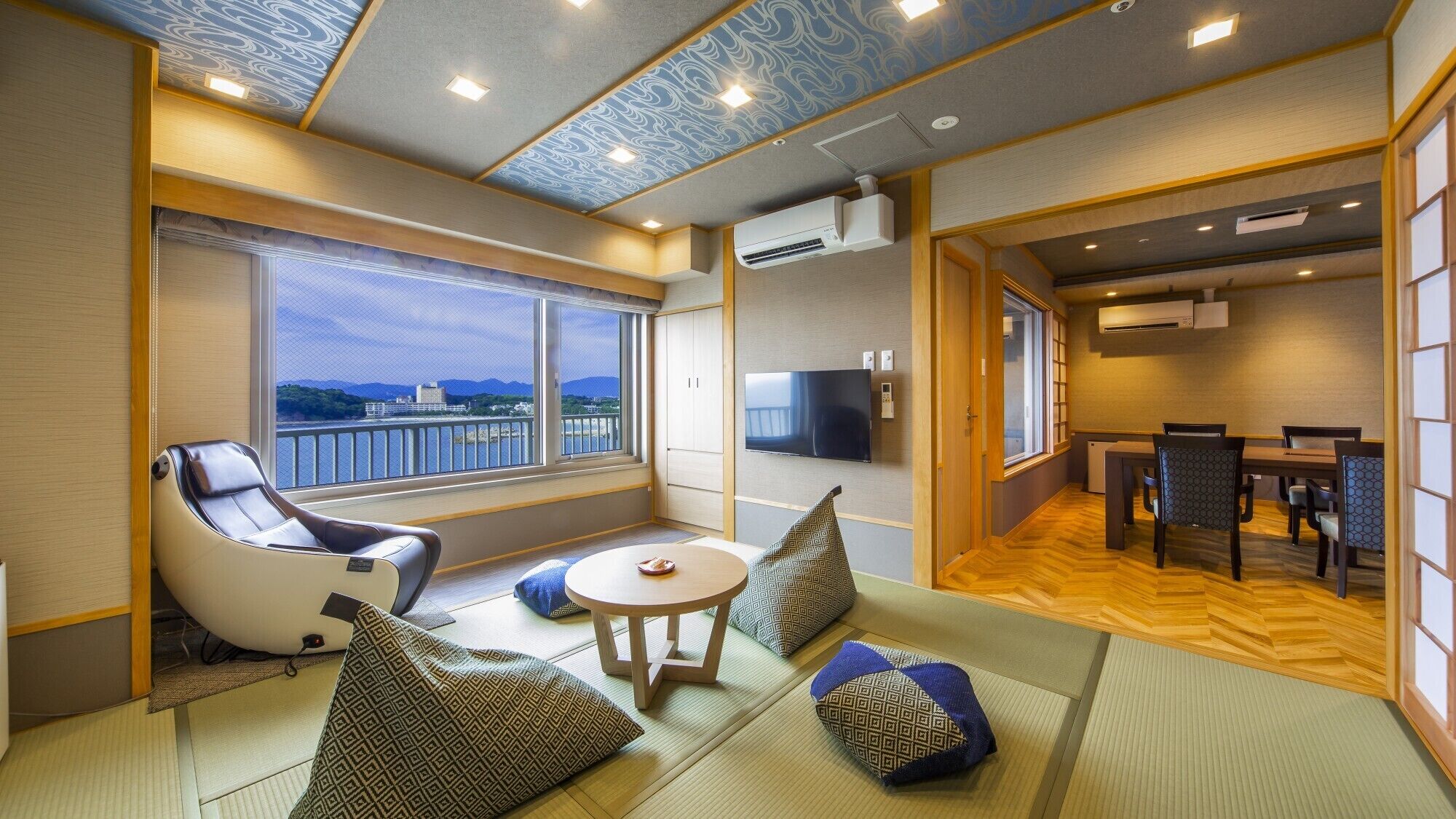 A room with tatami mats, a massage chair, a TV, and a view of the city.