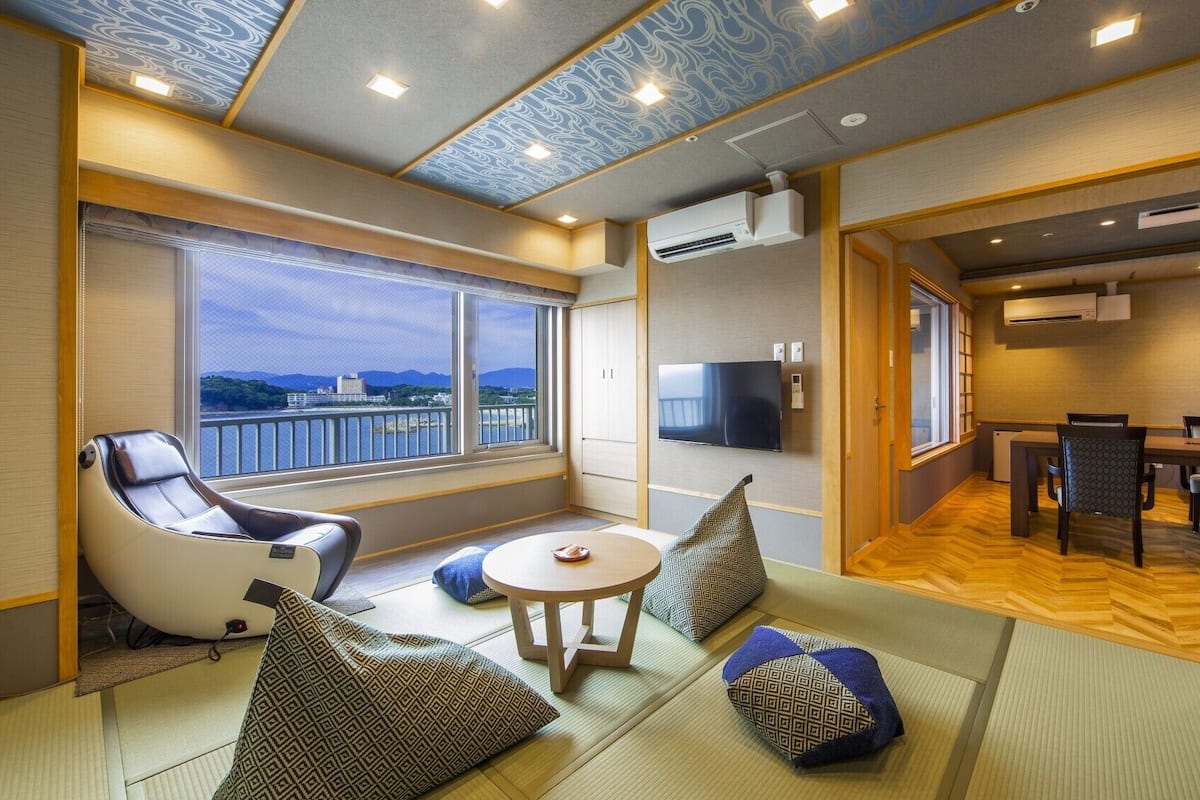 A room with tatami mats, a massage chair, a TV, and a view of the city.