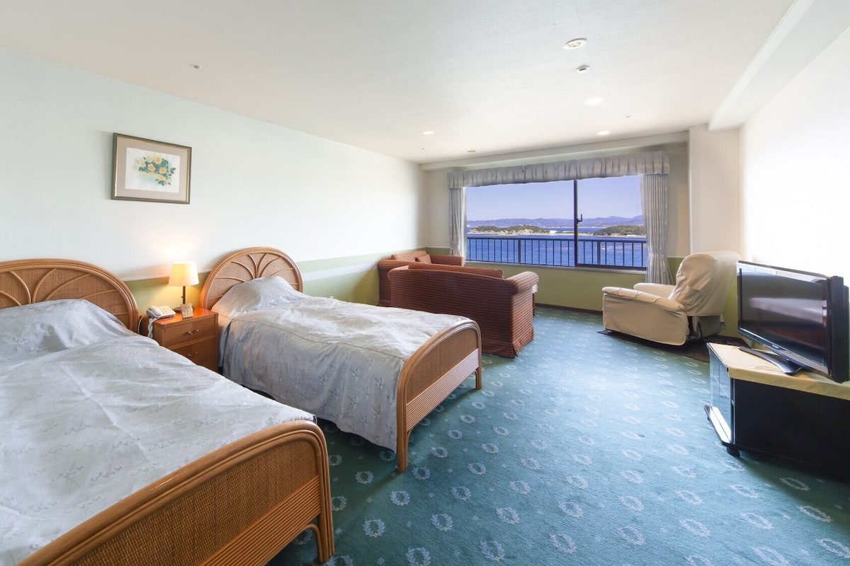 A hotel room with two beds, a television, a chair, and a view of the sea.