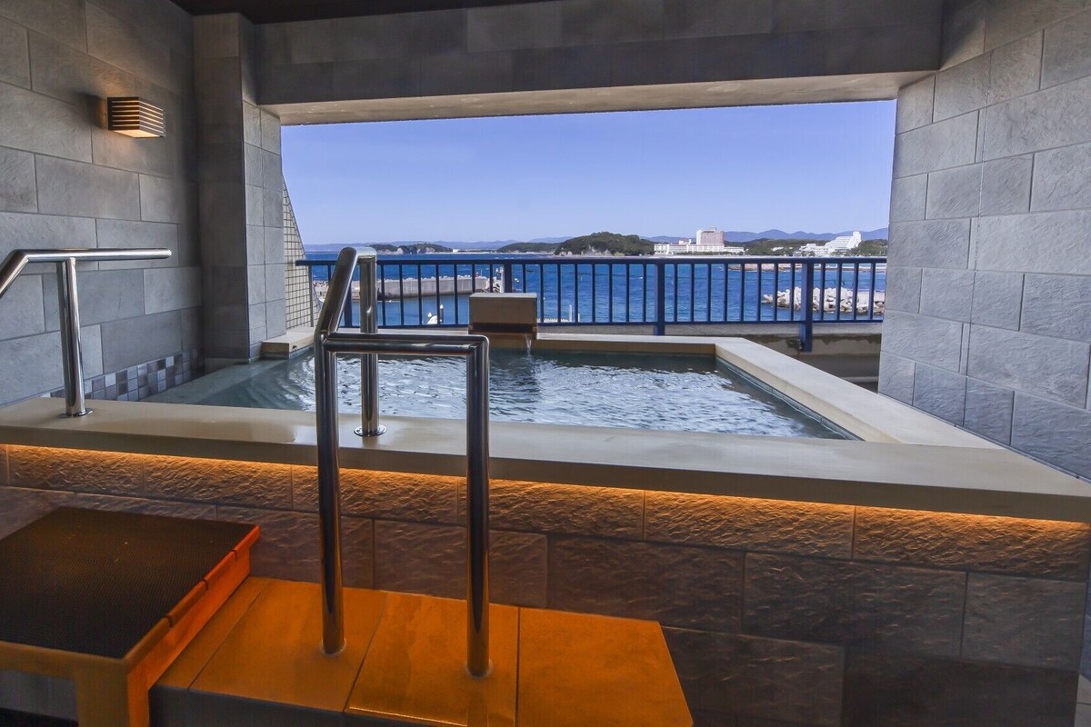 A modern indoor pool with a view of the sea, a metal staircase, and a stone wall.