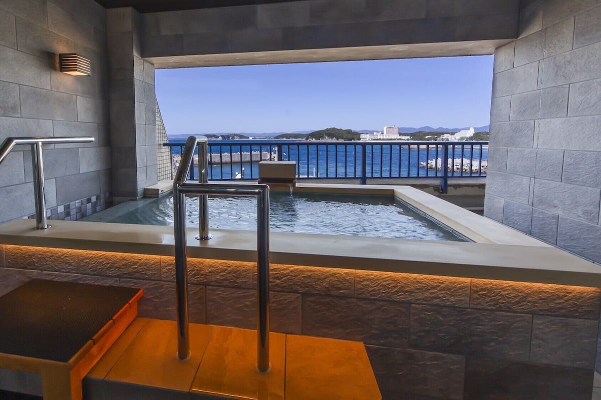 A modern indoor pool area with a view of the sea, a metal staircase, and a stone wall.