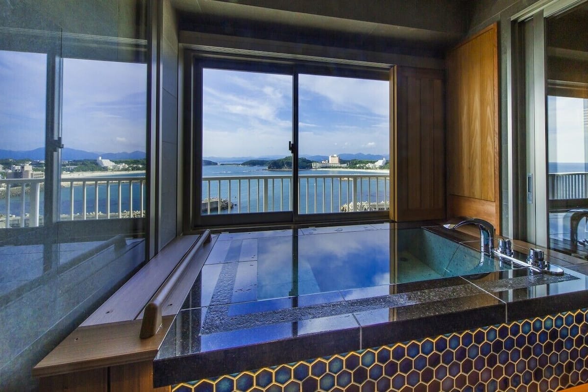 A modern bathroom with a large window, a glass-enclosed bathtub, and a view of a coastal area.