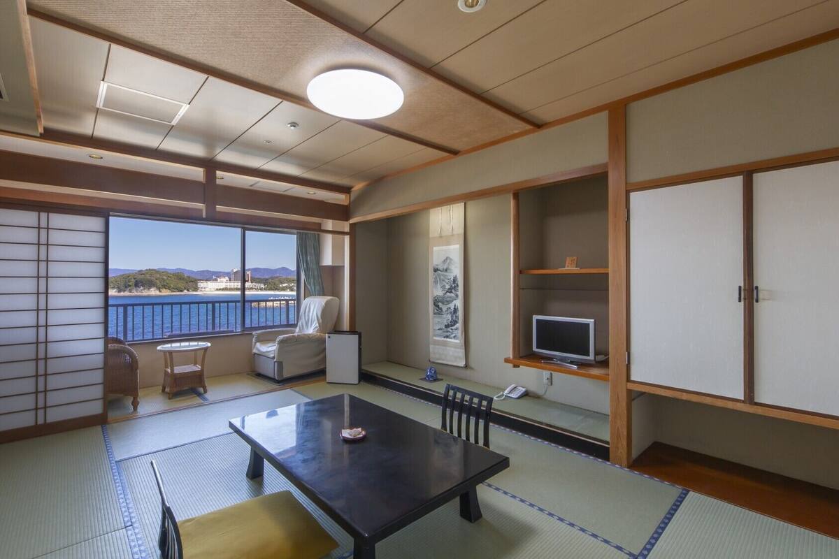 A room with tatami flooring, a tatami mat table, a sofa, a TV, and a view of the sea.