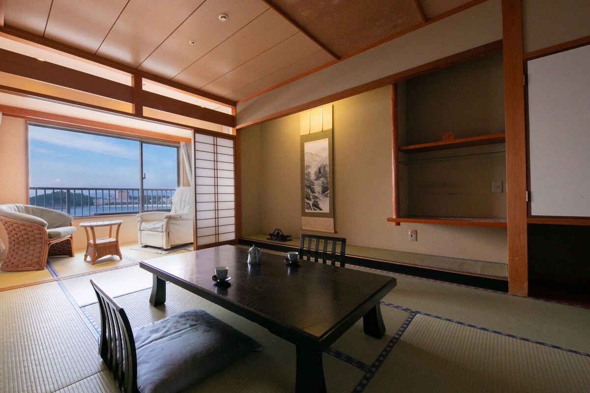 A traditional Japanese room with tatami flooring, a low table with cushions, a sofa, and a sliding door leading to a balcony with a view.