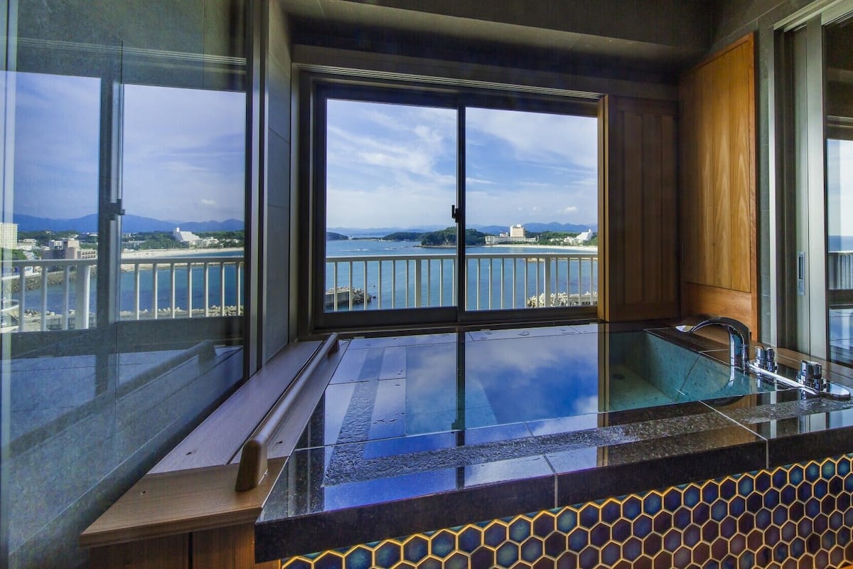 A modern bathroom with a glass-enclosed bathtub, wooden paneling, and a view of a coastal area.