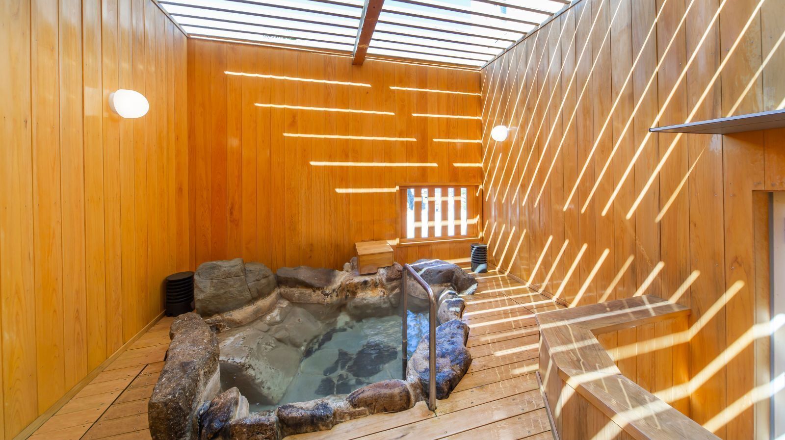 A wooden interior with a hot spring, wooden benches, and a stone wall.