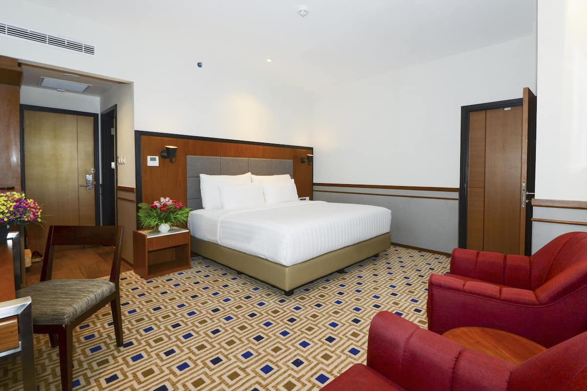 A hotel room with a bed, a desk with a chair, a red armchair, and a patterned carpet.