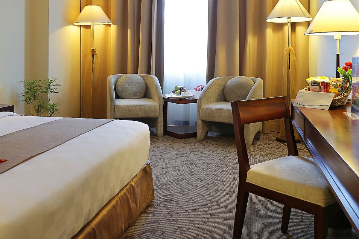 A hotel room with a large bed, two armchairs, a small table, and a desk with a lamp.
