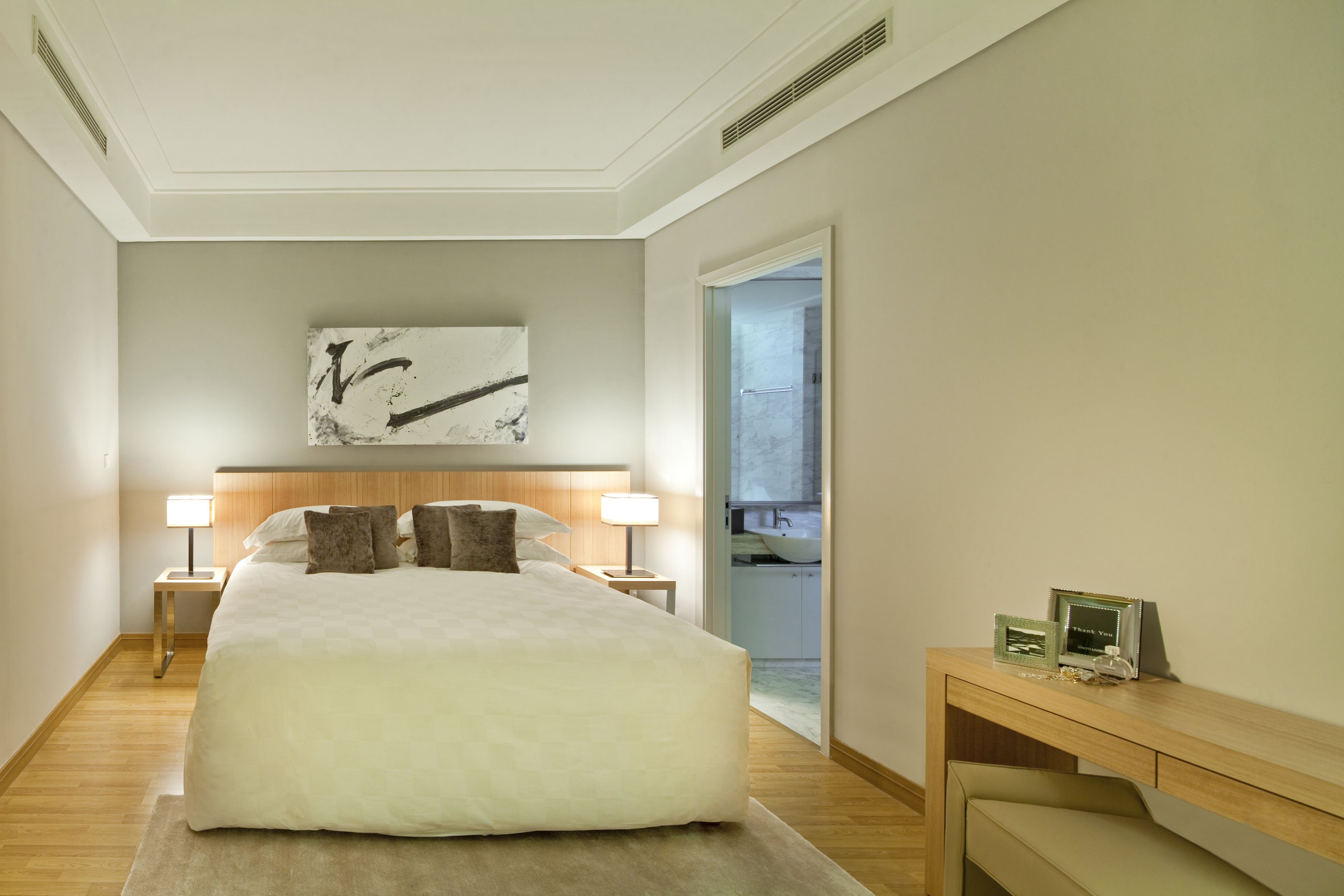 A hotel room with a large bed, two bedside tables with lamps, a wooden headboard, a wall-mounted artwork, and a view into the bathroom.