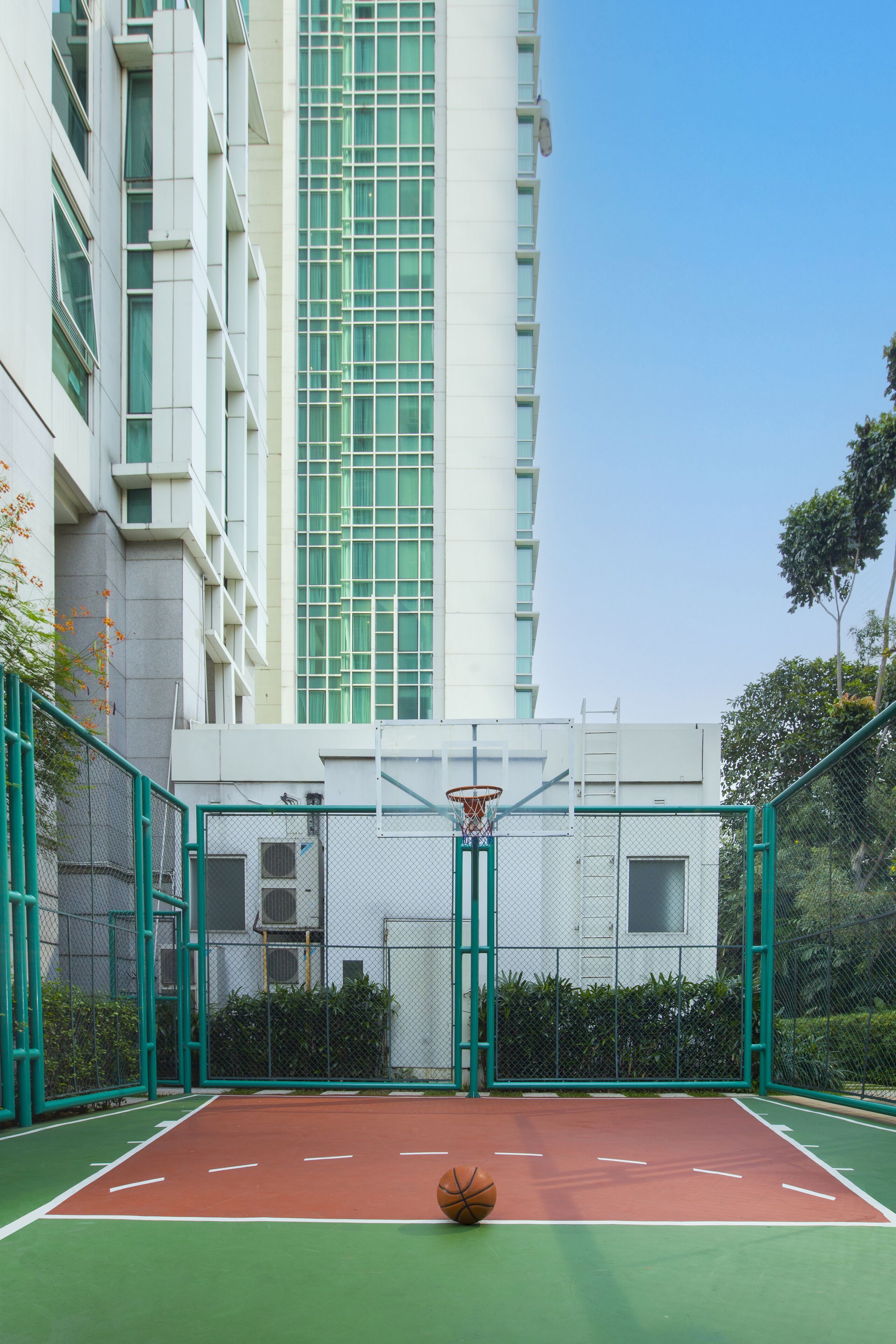 Una cancha de baloncesto al aire libre con aro, rodeada por una valla verde, y un edificio con grandes ventanales al fondo.
