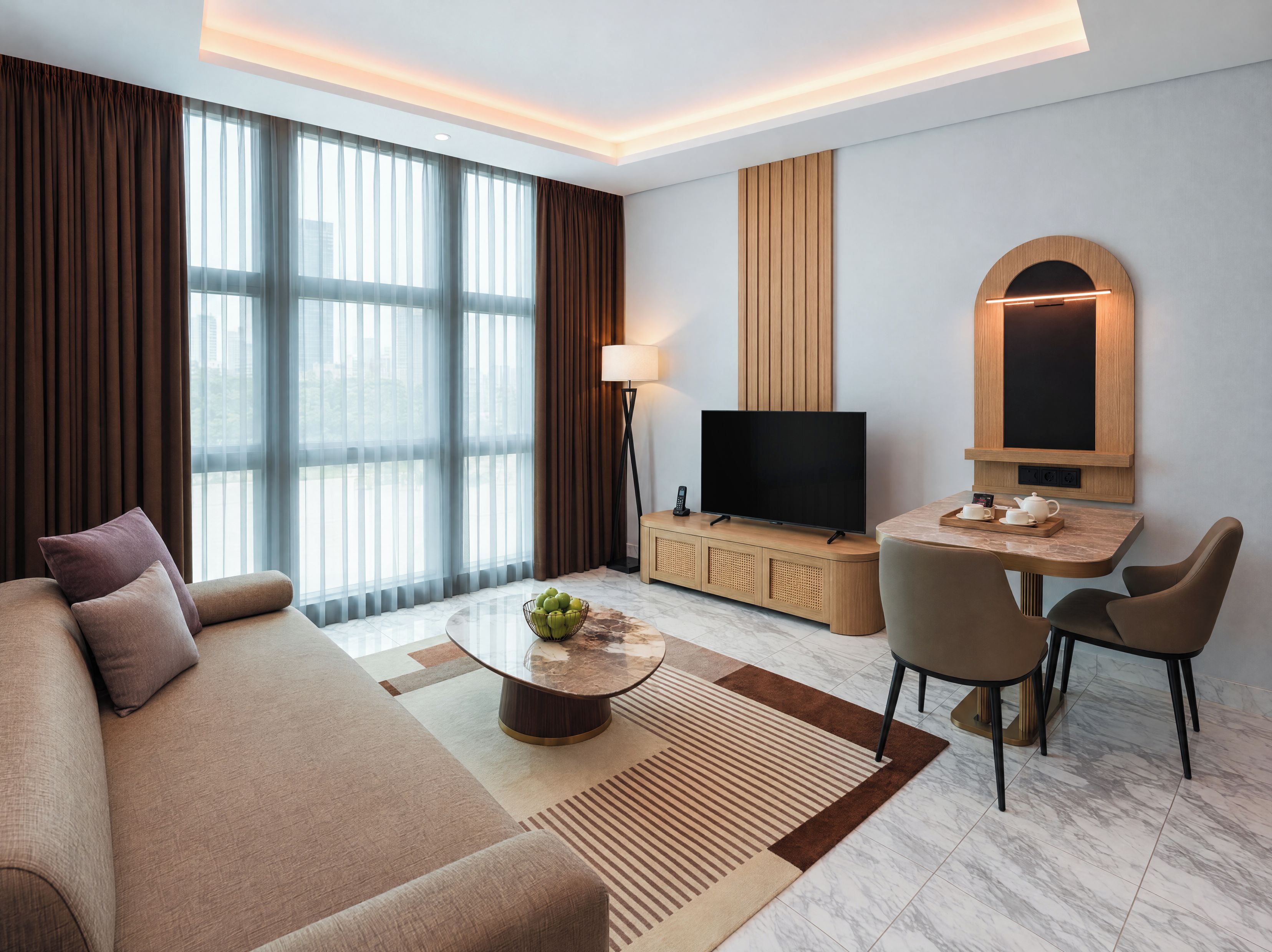 Una habitación de hotel moderna con sofá, una mesa de centro, televisión, comedor y ventanas amplias.