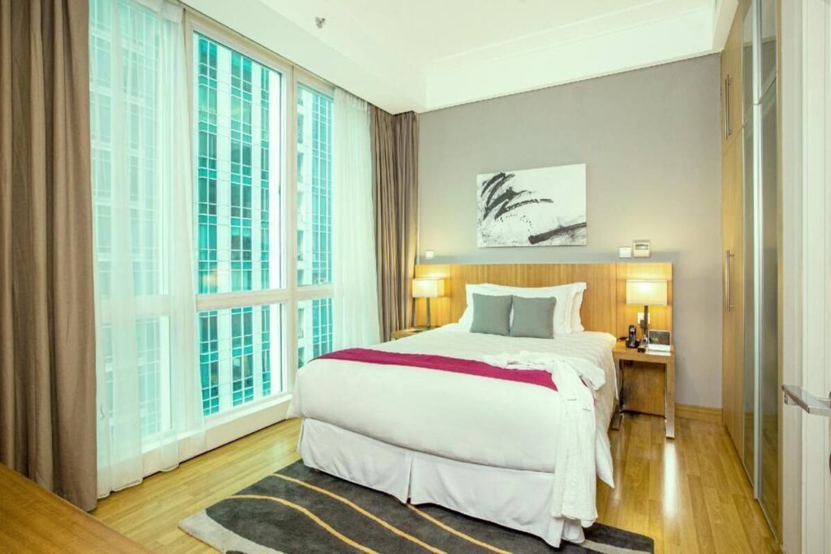 A hotel room with a large window, a bed with white bedding, two bedside lamps, a wooden headboard, and a painting on the wall.