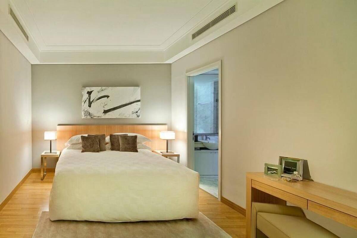 A hotel room with a large bed, two bedside tables with lamps, a wooden headboard, a wall art piece, and a desk with a chair.