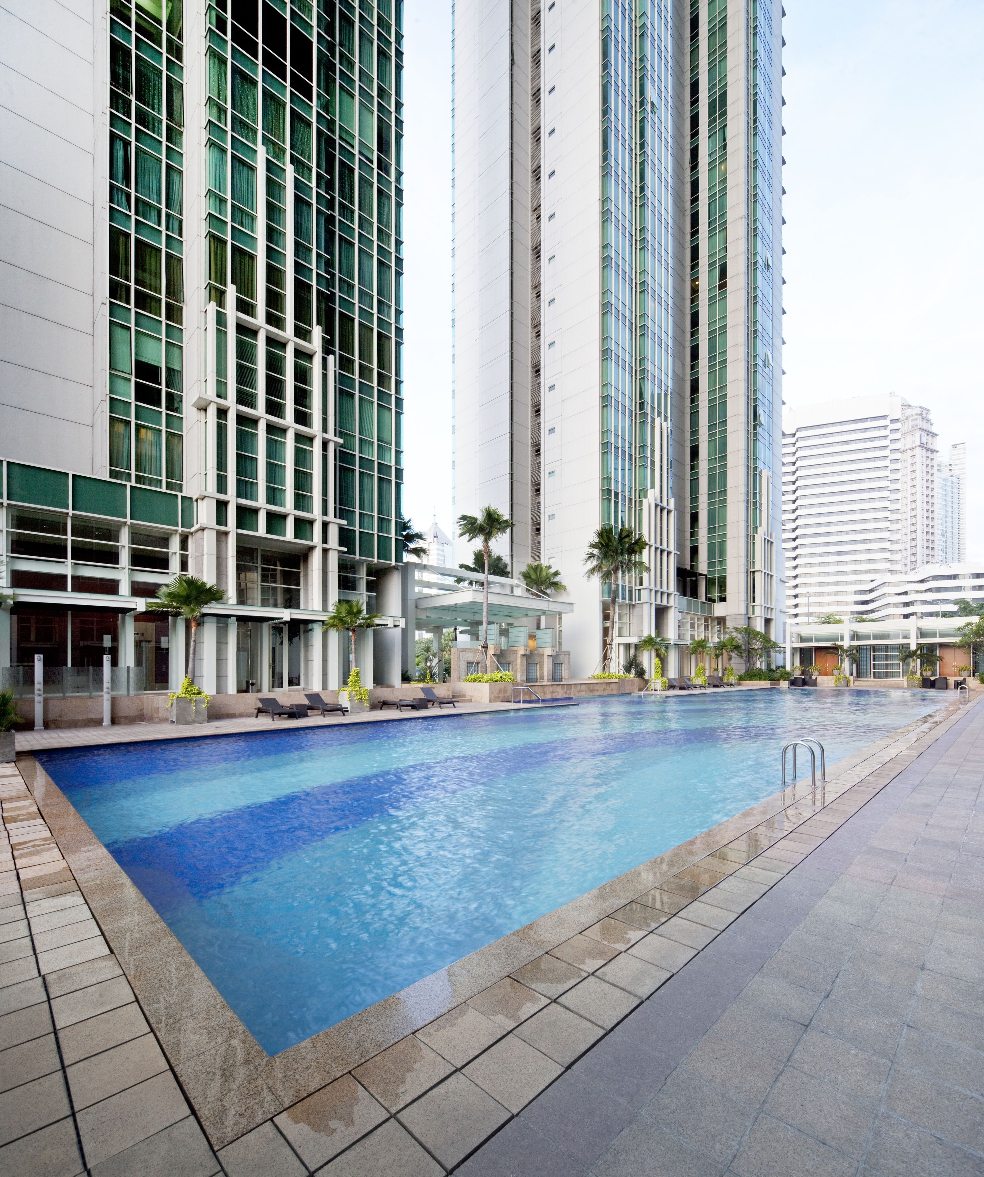 A swimming pool with a tiled deck, surrounded by tall buildings with large windows and palm trees.