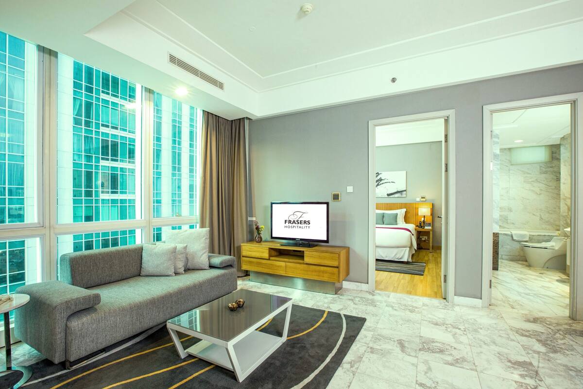 A modern hotel room with a grey sofa, a flat-screen TV, a wooden console, a large window, and a bathroom visible through an open door.